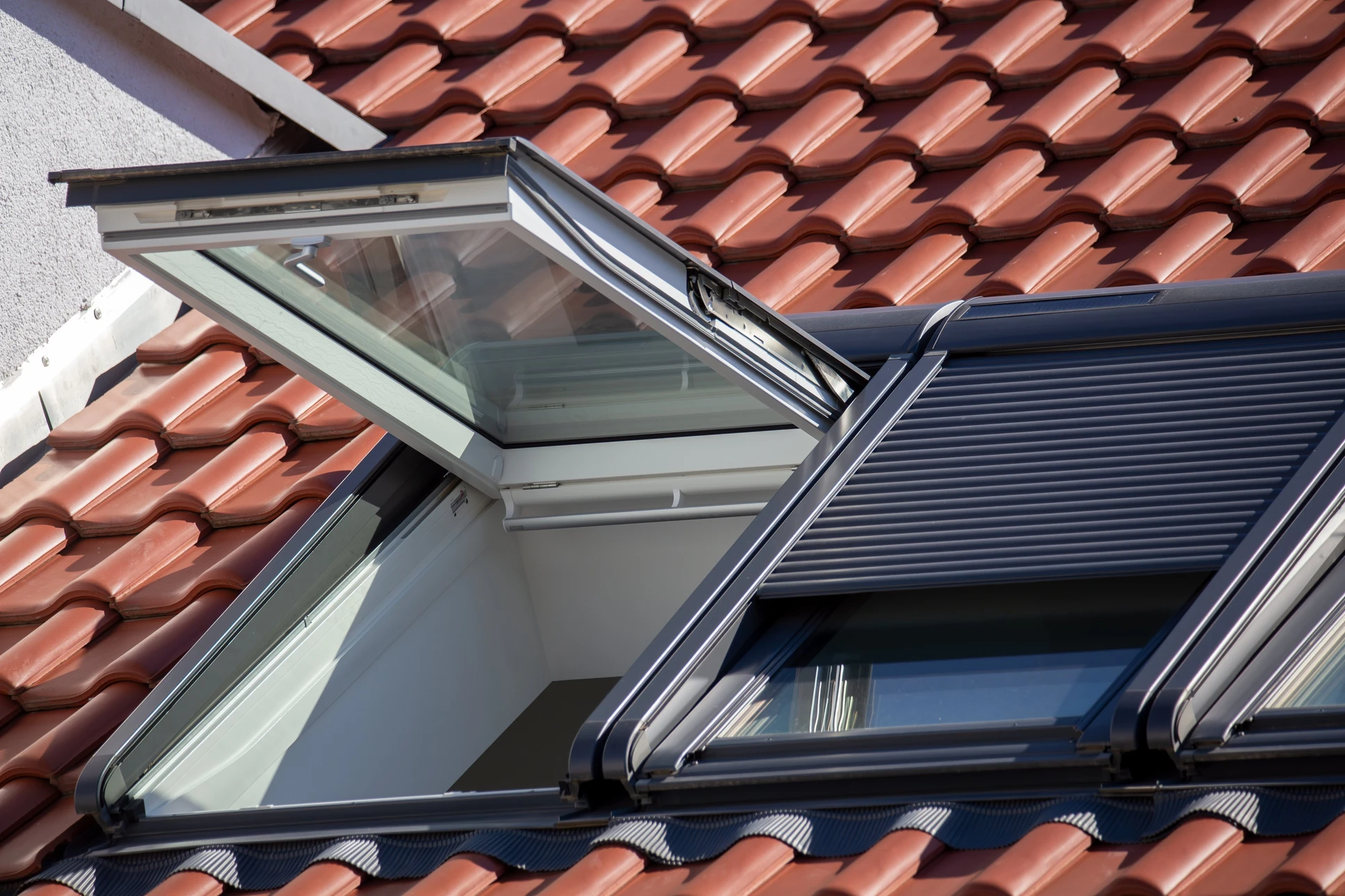Gros plan d'un volet roulant solaire Vélux en aluminium, entièrement déployé, monté sur une fenêtre de toit dans une pièce mansardée lumineuse. Le volet doit montrer les lamelles de la toile roulante, la barre inférieure, les rails de guidage latéraux. La lumière extérieure doit bien montrer le contraste : pièce très sombre côté volet déployé (obscurité totale) et lumière naturelle visible à travers les parties non occultées de la fenêtre. Ambiance intérieure de chambre ou séjour confortable sous pente.